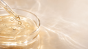 Pipetta di vetro che rilascia una goccia di siero alla niacinamide in una capsula Petri, macro scientifica, Chrissie Cosmetics