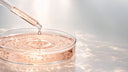 Pipetta di vetro da laboratorio che rilascia una goccia di fluido color pesca chiaro in una capsula Petri, con bolle e strutture molecolari sospese nel liquido, Chrissie Cosmetics