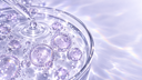 Pipetta di vetro che rilascia una goccia in una capsula Petri con vescicole liposomiali traslucide dai riflessi lilla, Chrissie Cosmetics